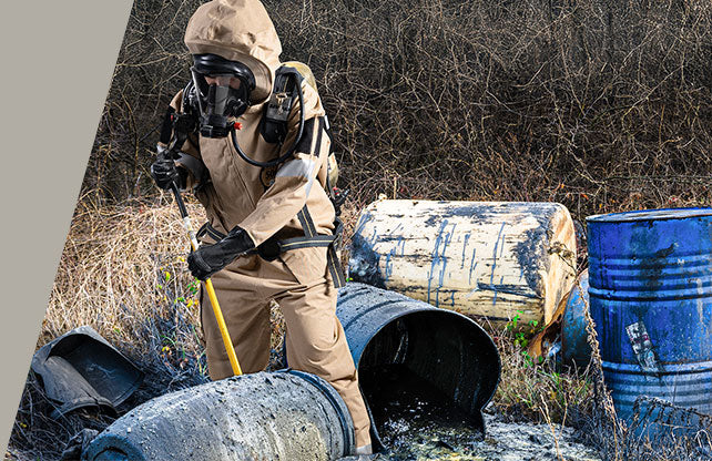 Person in hazmat suit and gas mask near toxic barrels in an outdoor setting